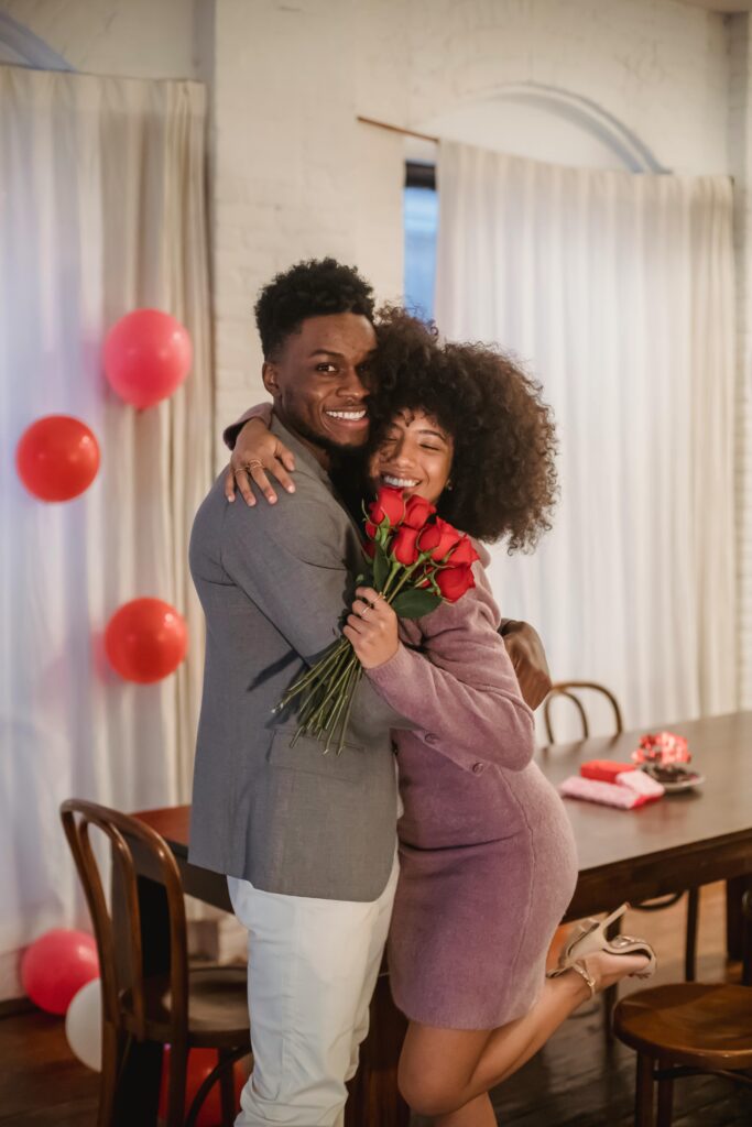 pexels photo 6579094 6579094 Cheerful African American couple with roses hugging and looking at camera while standing in room decorated with balloons during holiday celebration